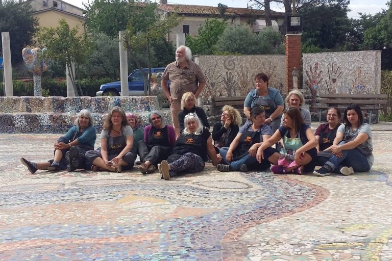 Gruppenfoto auf dem Mosaikplatz Ovales Foto einer Gruppe von Personen, die auf einem großen, kunstvollen Mosaikboden im Freien sitzen, mit einer verzierten Mosaikwand im Hi