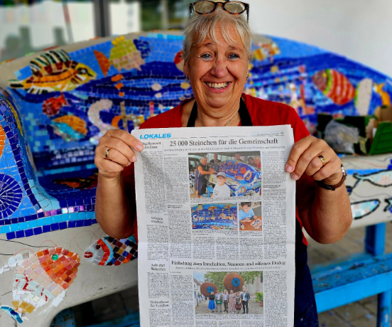 Pressebericht für das Mosaikprojekt '25 000 Steinchen' Eine lächelnde Mosaikkünstlerin hält eine Zeitung mit dem Artikel '25 000 Steinchen für die Gemeinschaft' vor einem großen blau-weißen Fisch