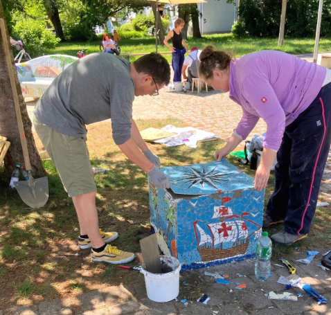 Gemeinschaftliches Mosaik-Projekt Engagierte Helfer und Künstler arbeiten gemeinsam an einem blauen Mosaik-Würfel mit einem Segelschiff-Motiv im Park.
