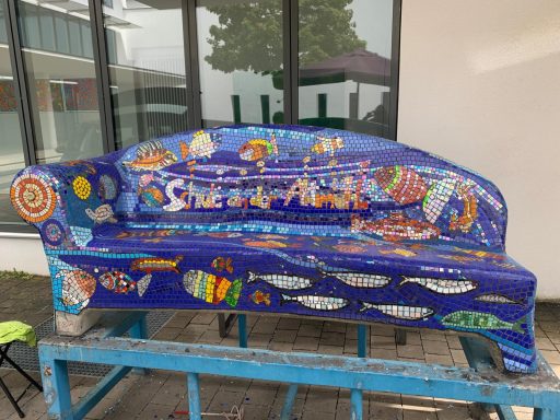 Mosaik-Sofa "Küstenstadt" Großes Mosaik-Sofa im Freien mit blauem Sitz, rot-gelber Armlehne und stilisierter Häuserlandschaft auf der Rückenlehne.