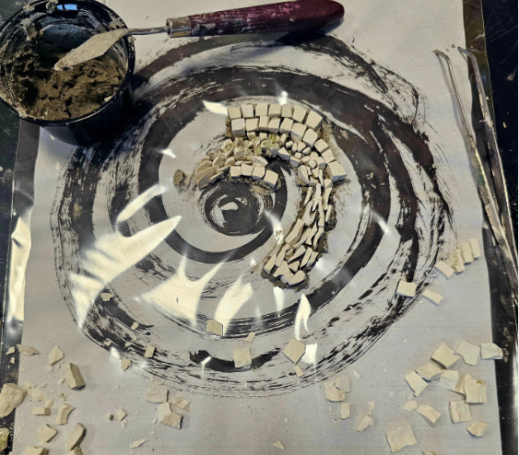 Entstehung eines Mosaiks: Helle quadratische Steine werden in einer Spiralform auf einem schwarz-weiß gemusterten Untergrund angeordnet.