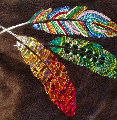 Glänzende Mosaik-Federn Drei kunstvolle, bunte Federn aus Mosaiksteinen und Perlen auf dunklem Untergrund.