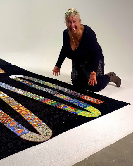 Caroline Jung beim Mosaik-Aufbau Künstlerin Caroline Jung sitzt am Boden neben dem wachsenden Mosaik-Band und erklärt den Aufbauprozess.