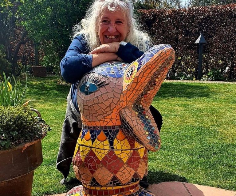 Lächelnde Frau stützt sich auf eine große, farbenfrohe Mosaik-Entenskulptur in einem sonnigen Garten.