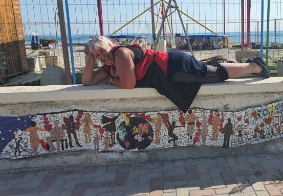 Frau in Schwarz und Rot posiert entspannt liegend auf der Mauerkrone, direkt über dem langen Wandmosaik mit Menschen und Erdkugel.