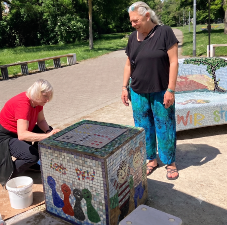 Künstlerische Arbeit im Freien Mosaikkünstlerin Caroline Jung und eine Helferin arbeiten im Freien an einem bunten Sitzwürfel mit Tiermotiven.