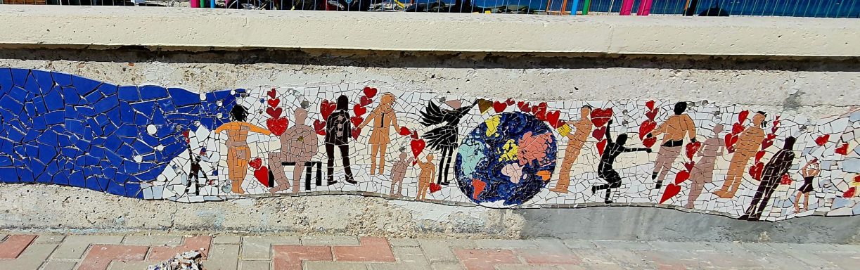 Detailaufnahme eines Wandmosaiks, das verbundene Menschenfiguren, rote Herzen, einen schwarzen Vogel und eine bunte Erdkugel auf weißem Grun