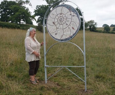 Mosaik-Skulptur "Ammonit" Große kreisförmige graue Mosaik-Skulptur mit Spiralmuster, montiert auf einem Metallgestell im Freien.