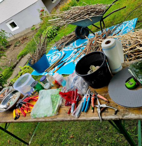 Ein Arbeitsplatz im Garten mit einer Schubkarre voller Zweige, Werkzeugen und Materialien, die für ein Kunstprojekt auf einer blauen Plane b