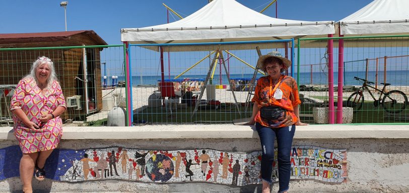Frau in orangefarbenem Oberteil steht neben einem langen Wandmosaik mit Menschenfiguren und Erdkugel an einer Strandpromenade.
