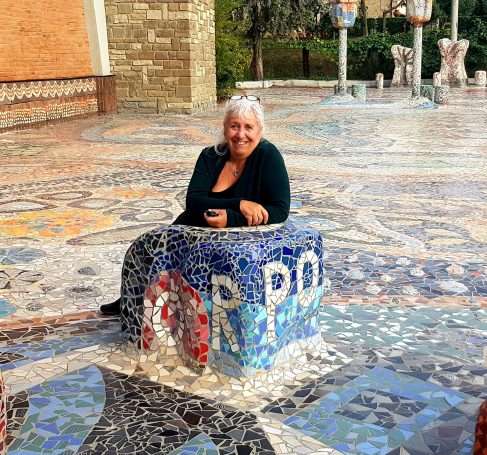 Lächelnde Frau mit hellen Haaren sitzt im Freien auf einem mit Mosaik verzierten Steinblock inmitten eines großen Mosaikplatzes.