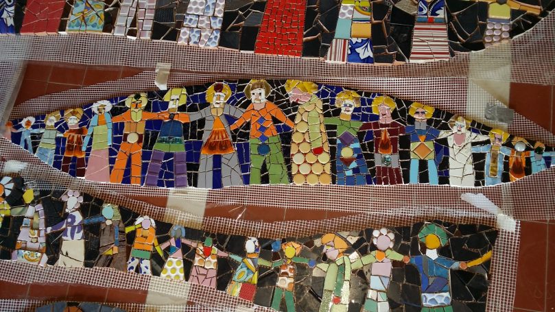 Großes Wandmosaik mit mehreren horizontalen Reihen von bunten Menschenfiguren, die sich an den Händen halten, abgetrennt durch dekorative St