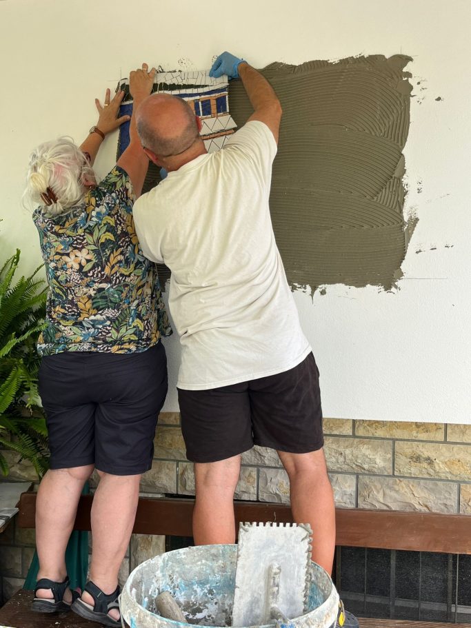 Mosaikkünstlerin Caroline Jung und ein Helfer bei der fachgerechten Montage eines großen Wandmosaiks auf einer vorbereiteten Fläche.