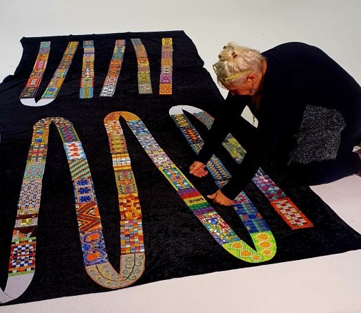 Montage des textilen Mosaik-Bandes Mosaikkünstlerin Caroline Jung kniet auf einem großen schwarzen Untergrund und ordnet Mosaikfragmente zu einem fortlaufenden Band an.