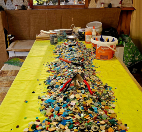 Ein Arbeitstisch mit einer gelben Tischdecke, darauf liegt ein langer Haufen loser Mosaiksteine und Werkzeuge wie Mosaikzangen.