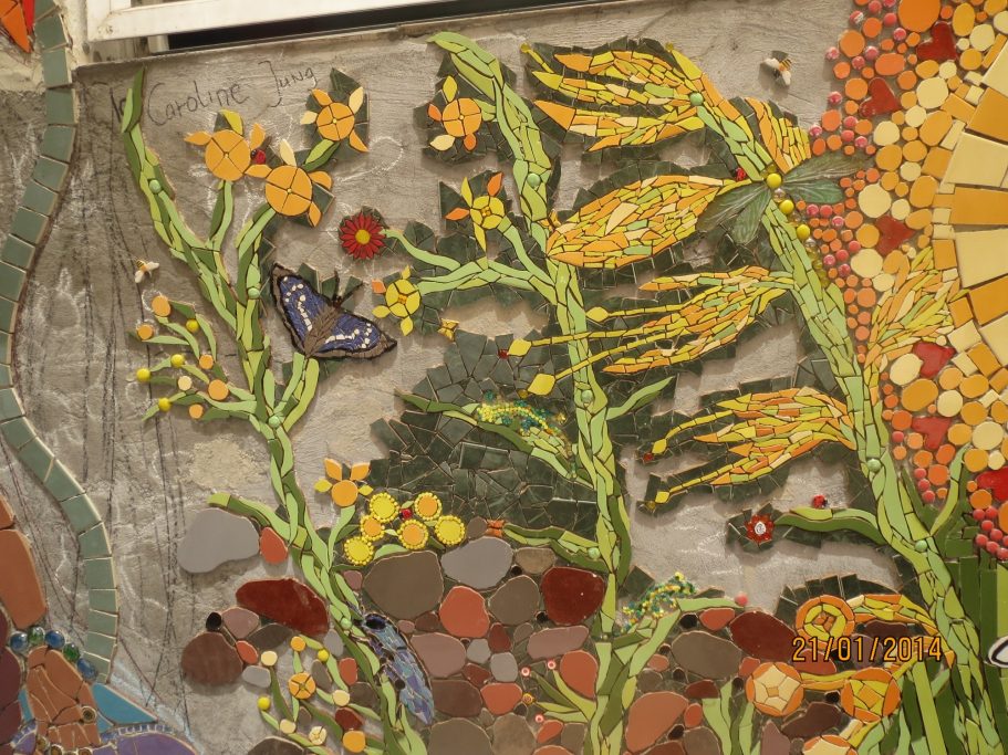 Detailaufnahme eines Wandmosaiks, das grüne Pflanzen, gelbe Blüten, braune Steine und einen Schmetterling zeigt.