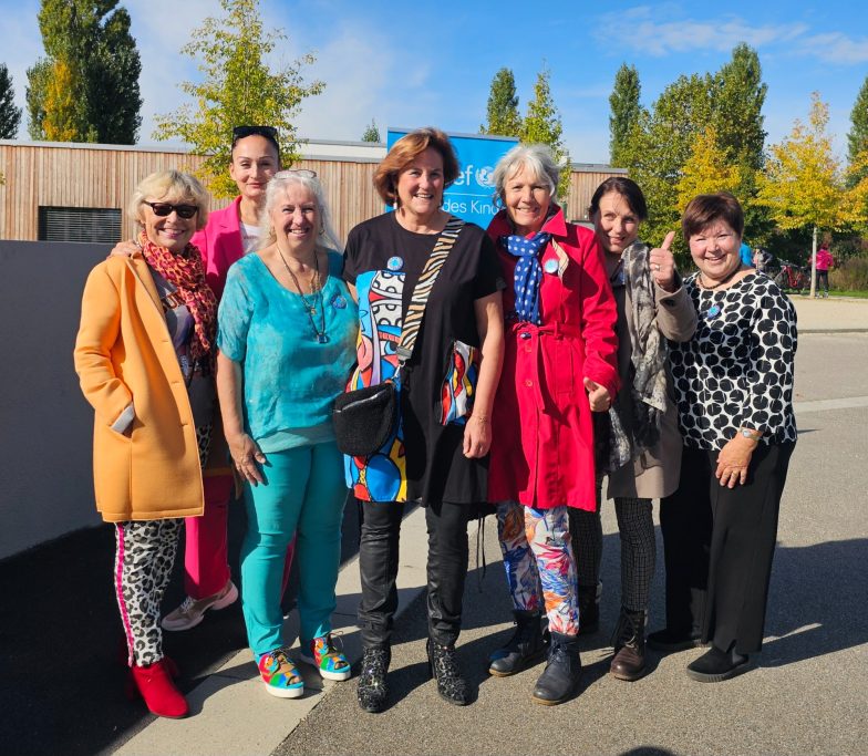 Das engagierte Team hinter dem Projekt Eine engagierte Gruppe von Frauen und Künstlerinnen posiert lächelnd vor einer bunten Wandgestaltung auf dem Schulhof.