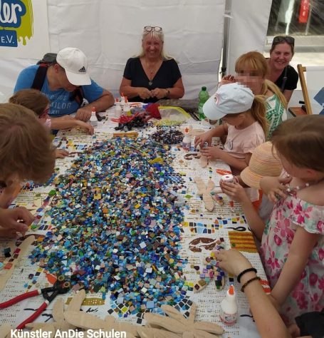 Erwachsene und Kinder arbeiten gemeinsam an einem großen Mosaikprojekt an einem Tisch im Freien, in der Mitte liegt ein riesiger Haufen blau