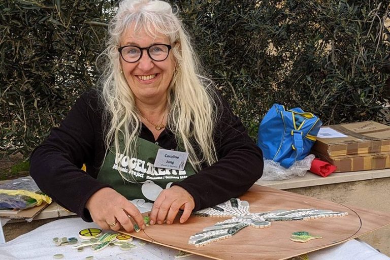 Caroline Jung arbeitet lächelnd in ihrem Atelier an der handwerklichen Umsetzung eines neuen Mosaik-Kunstwerks.
