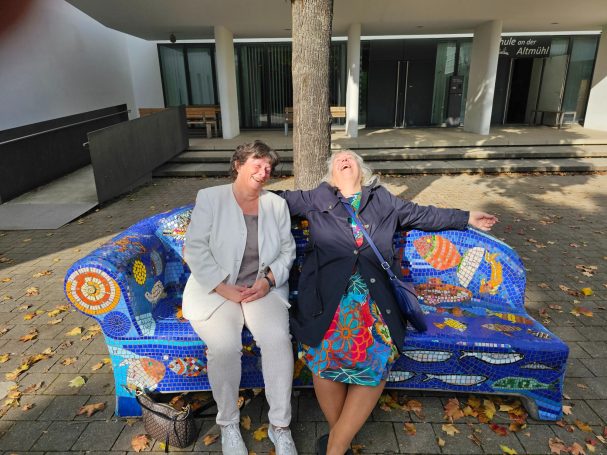 Freude auf dem Mosaik-Sofa Mosaikkünstlerin Caroline Jung und eine Begleiterin sitzen lachend auf einem großen, blauen Mosaik-Sofa im Freien.