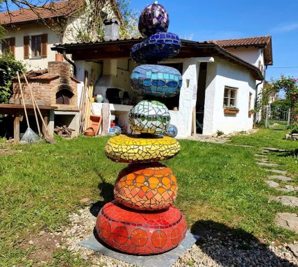 Stacked-Stone Mosaik-Totem Hohe Mosaik-Totempfahl-Gartenskulptur aus gestapelten bunten kreisförmigen Steinen vor einem rustikalen Gebäude.