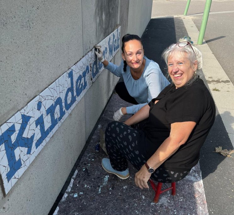 Gemeinschaftliches Mosaikprojekt an der Schule Mosaikkünstlerin Caroline Jung und eine Kollegin arbeiten fröhlich an einem blauen Mosaik-Schriftzug an einer Schulwand in Ingolstadt.