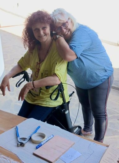 Zwei lachende Frauen in einer Werkstatt umarmen sich an einem Tisch mit Mosaikwerkzeugen; eine sitzt neben einem Rollator.