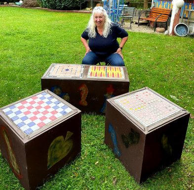 Mosaik-Schachtische im Garten Frau sitzt im Grünen neben drei dunklen Tischen mit Mosaik-Schachbrettmustern auf der Platte.