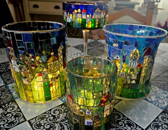 Mosaik-Windlichter "Stadtlandschaft" Gruppe von mehreren beleuchteten Mosaik-Gläsern mit farbenfrohen Stadt- und Häusermotiven auf einem gemusterten Tisch