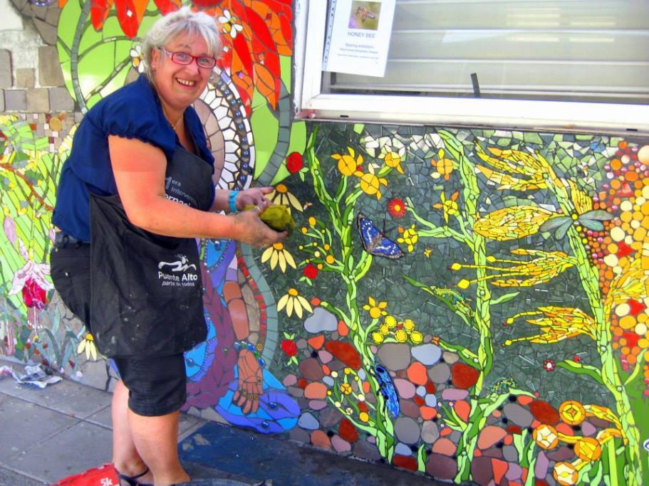 Eine lächelnde ältere Frau in einer schwarzen Schürze arbeitet an einem farbenprächtigen Wandmosaik mit floralen Mustern.