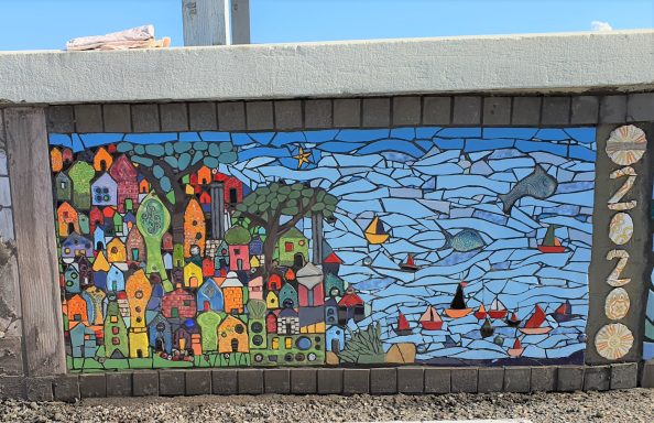 Buntes Mosaik-Wandbild einer stilisierten Küstenstadt mit bunten Häusern, Bäumen und einem Meer voller Fische und Boote.