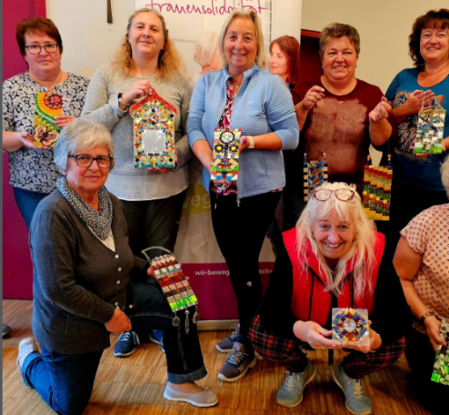Eine Gruppe von Frauen präsentiert drinnen freudig ihre selbstgemachten, bunten Mosaik-Kunstwerke, die wie kleine Häuser aussehen.