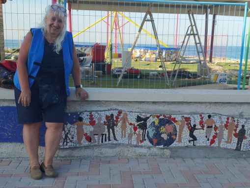 Ältere Frau im blauen Oberteil steht stolz neben dem fertiggestellten Mosaikband an der Mauer vor einem Spielplatz.