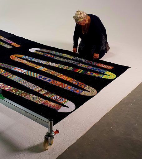 Das fortlaufende Mosaik-Kunstwerk Weitwinkelaufnahme des fast fertigen, schlangenförmigen Mosaik-Bandes auf schwarzem Untergrund, an dem Caroline Jung arbeitet.