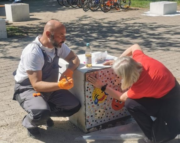 Mann und Frau arbeiten gemeinsam an einem großen Mosaikwürfel im Freien.